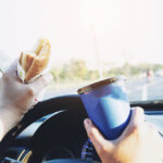 Distracted driver in a vehicle with a blue travel mug and sandwich in hand