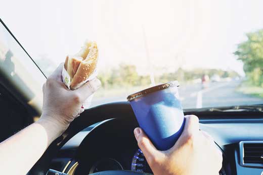Distracted driver in a vehicle with a blue travel mug and sandwich in hand
