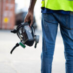 Person in neon green shirt and blue jeans stands holding a gas mask