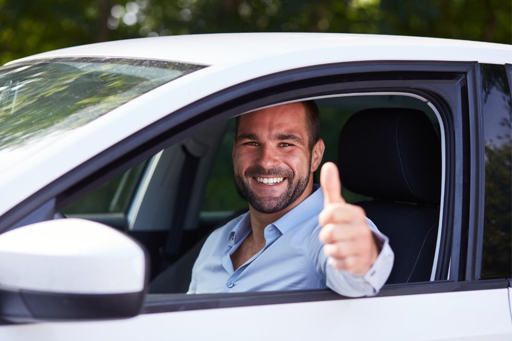 man sits in front seat of car giving thumbs up to camera