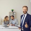 smiling insurance broker gives a thumbs up to the camera with clients in background