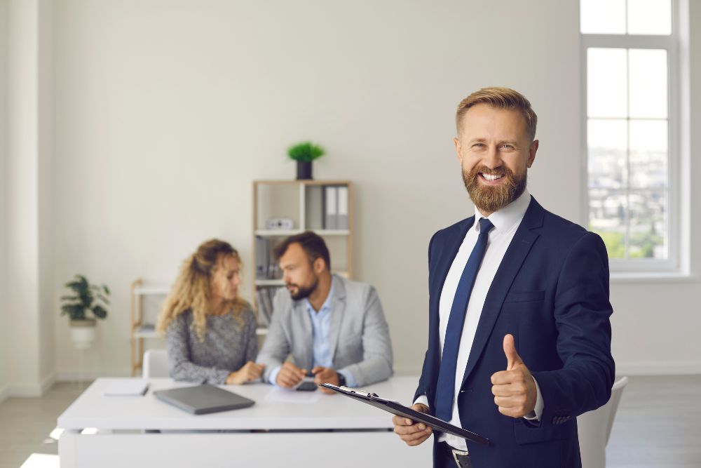 smiling insurance broker gives a thumbs up to the camera with clients in background