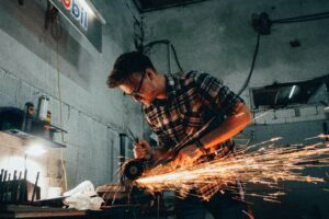 Man in plaid shirt using angle grinder on a piece of metal in dark shop