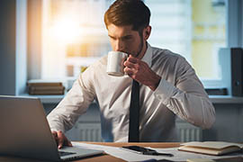 Man in white shirt and black tie sits at a wooden desk, drinking coffee and working on a laptop