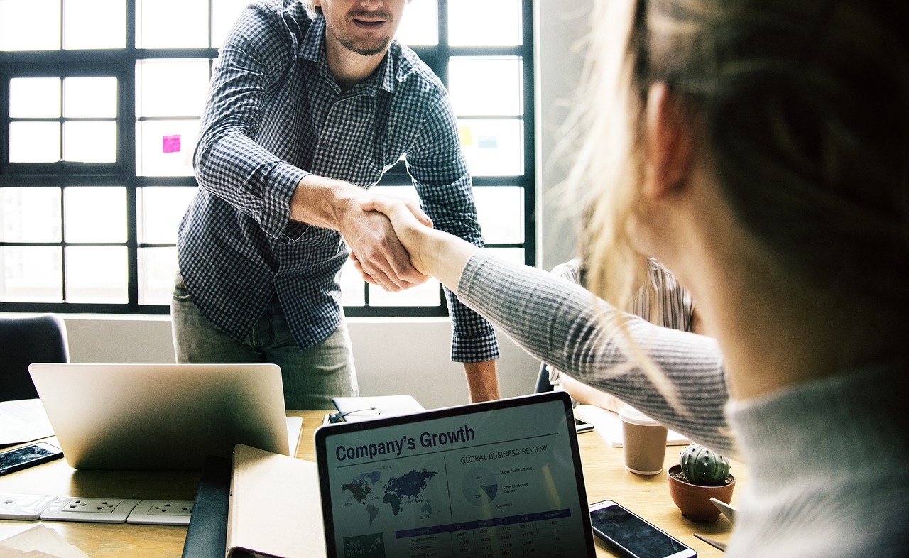 Man in checkered shirt and woman in a grey sweater shake hands over a wooden desk. Both have laptops, one displaying a company growth chart.